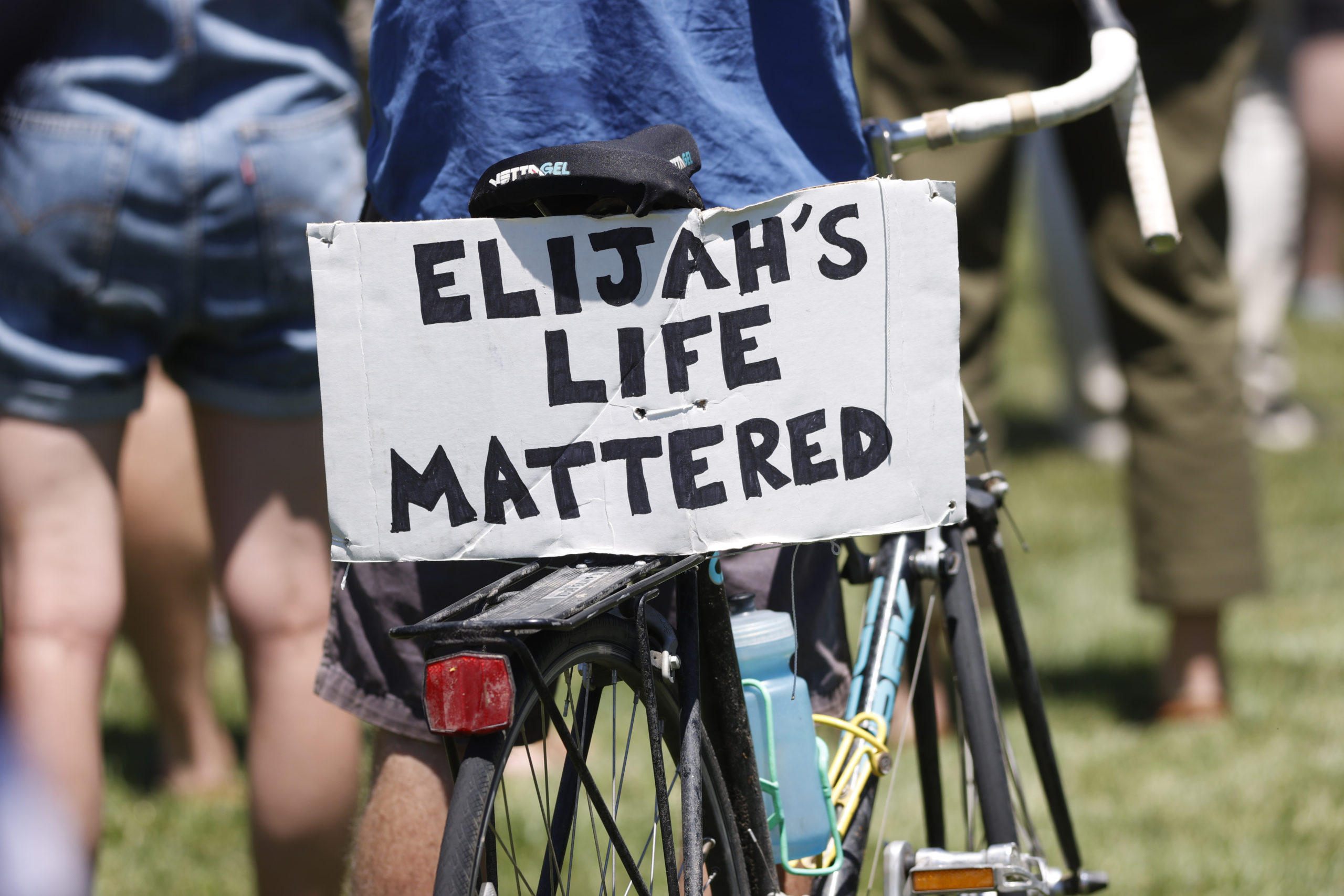 A placard is displayed on a bicycle during a rally and march over the death of 23-year-old Elijah McClain, Saturday, June 27, 2020, outside the police department in Aurora, Colorado. McClain died in late August 2019, after he was stopped while walking to his apartment by three Aurora Police Department officers. Photo credit: David Zalubowski, The Associated Press