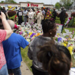 People gather around a memorial for the victims of the Buffalo supermarket shooting outside the Tops Friendly Market on Saturday, May 21, 2022, in Buffalo, N.Y. Tops was encouraging people to join its stores in a moment of silence to honor the shooting victims Saturday at 2:30 p.m., the approximate time of the attack a week earlier. Buffalo Mayor Byron Brown also called for 123 seconds of silence from 2:28 p.m. to 2:31 p.m., followed by the ringing of church bells 13 times throughout the city to honor the 10 people killed and three wounded. Photo credit: Joshua Bessex, The Associated Press