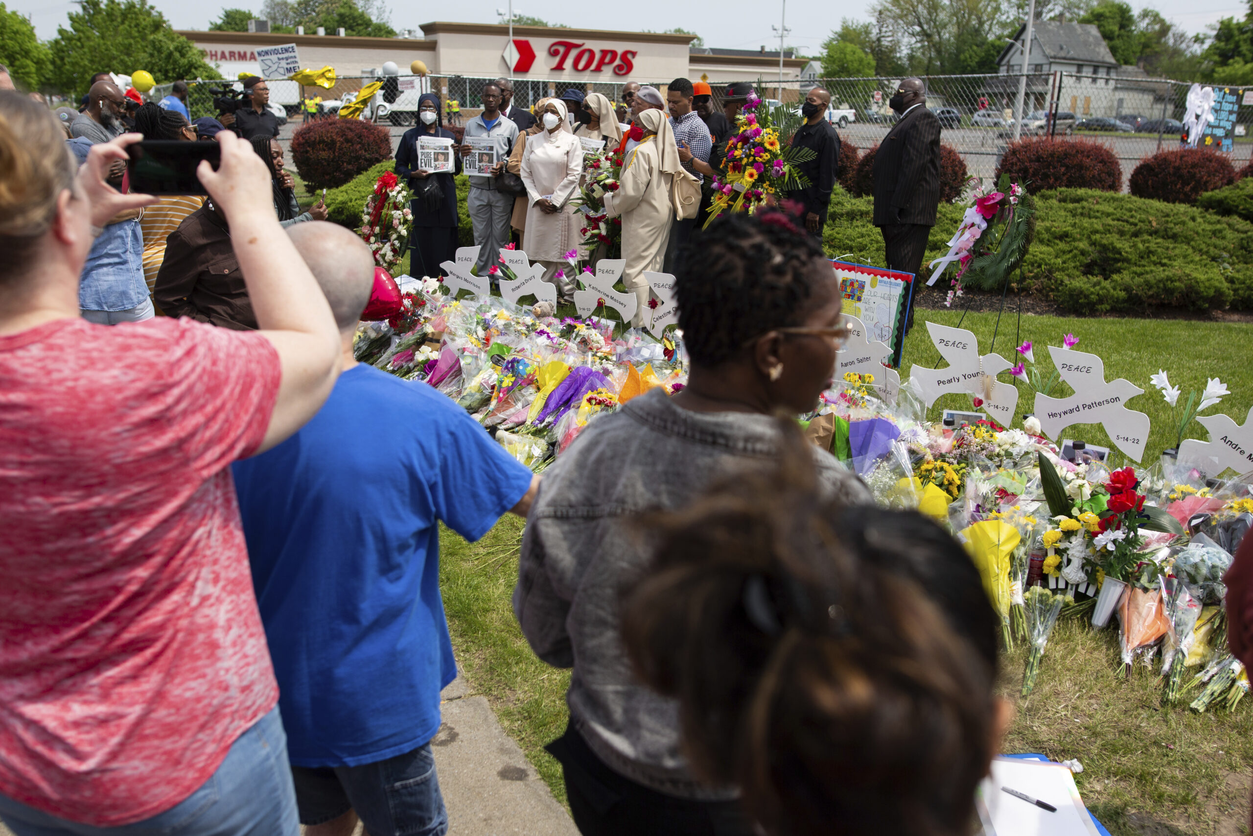 People gather around a memorial for the victims of the Buffalo supermarket shooting outside the Tops Friendly Market on Saturday, May 21, 2022, in Buffalo, N.Y. Tops was encouraging people to join its stores in a moment of silence to honor the shooting victims Saturday at 2:30 p.m., the approximate time of the attack a week earlier. Buffalo Mayor Byron Brown also called for 123 seconds of silence from 2:28 p.m. to 2:31 p.m., followed by the ringing of church bells 13 times throughout the city to honor the 10 people killed and three wounded. Photo credit: Joshua Bessex, The Associated Press
