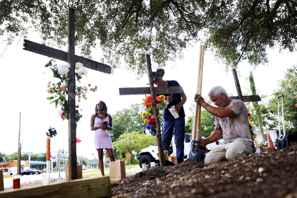 From left, a woman who goes by the name of "Queen" views crosses put up in memory of the victims of Saturday's shooting as artist Roberto Marquez, of Dallas, Texas, paints and Will Walsh, of Nocatee, Florida, helps construct posts Monday, Aug. 28, 2023, near the site of the attack at a Dollar General store in Jacksonville, Florida. Queen says she is a manager at the store and was holed up in the office at the store when the shooting occurred. Photo credit: Corey Perrine, The Florida Times-Union via The Associated Press