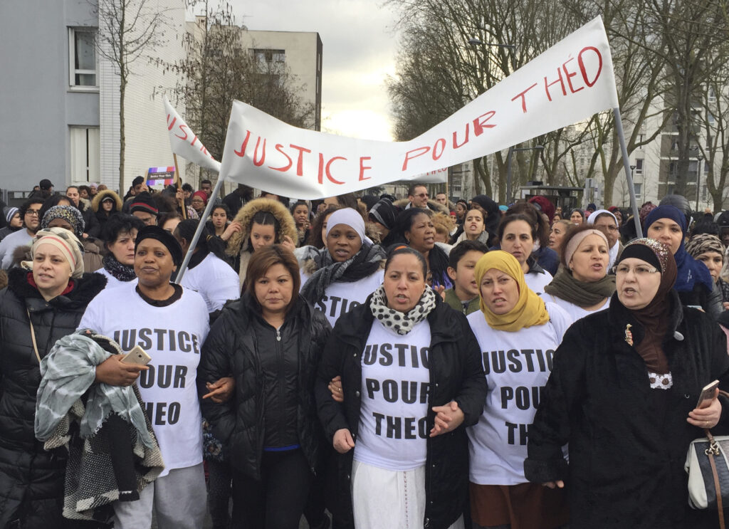 People march in the streets of Aulnay-sous-Bois, north of Paris, France, holding a sign reading "Justice for Theo" during a protest, a day after a French police officer was charged with with violence during an identity check, Monday, Feb. 6, 2017. A French court convicted three police officers of “voluntary violence” towards a youth worker in a Paris suburb who had a police baton forcibly inserted into his rectum during an identity check seven years ago. Photo credit: Milos Krivokapic, The Associated Press