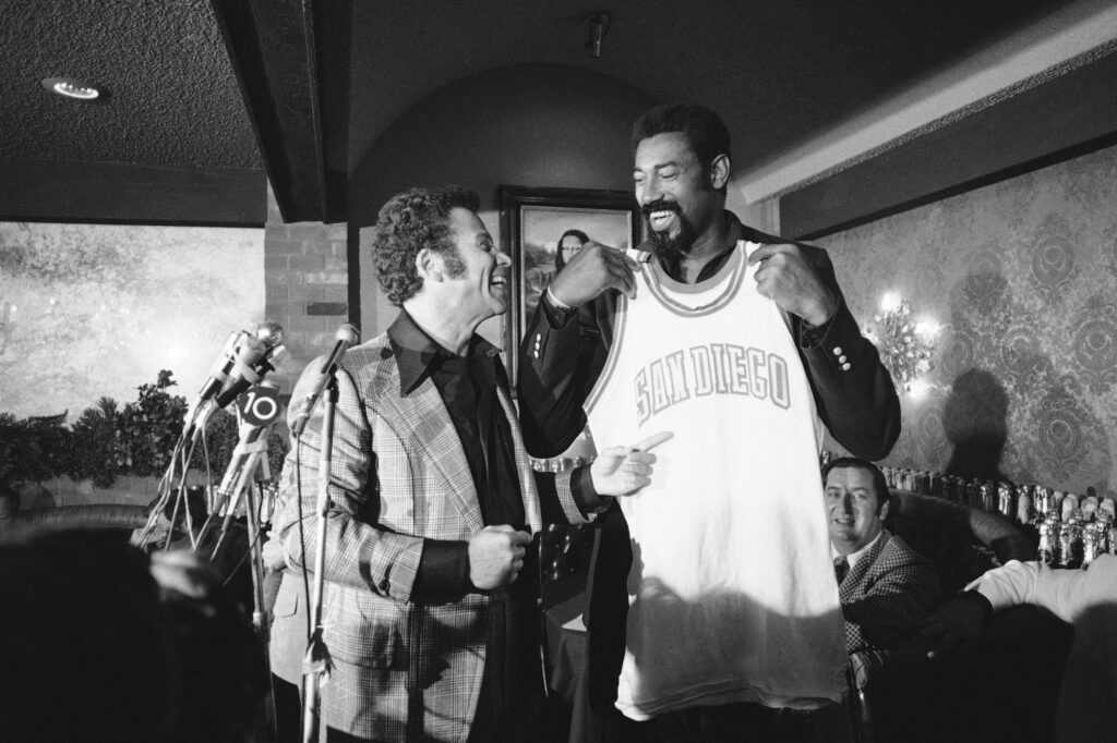Wilt Chamberlain, the veteran Los Angeles Lakers center, holds up his new shirt at news conference, Wednesday, Sept. 26, 1973 in Chula Vista, California, with Leonard Bloom, at left, owner of the San Diego Conquistadors. Chamberlain is joining the American Basketball Association team as player-coach. Photo credit: The Associated Press