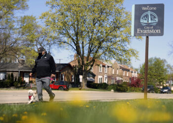 A person walks a dog past a street sign reading "Welcome to Evanston" in the predominantly Black 5th Ward in Evanston, Illinois, Tuesday, May 4, 2021. The Chicago suburb is preparing to pay reparations in the form of housing grants to Black residents who experienced housing discrimination. ​Photo credi: Shafkat Anowar, The Associated Press