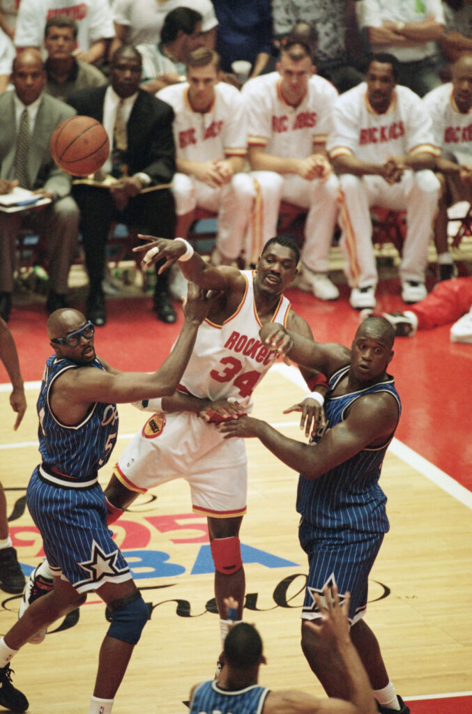 Then-Houston Rockets center Hakeem Olajuwon (34) passes the ball as he is pressured by Then-Orlando Magic players Shaquille O'Neal (32) and Horace Grant (54) during the second quarter of Game 3 of the NBA Finals at Houston on June 11, 1995. Photo credit: Rick Bowmer, The Associated Press
