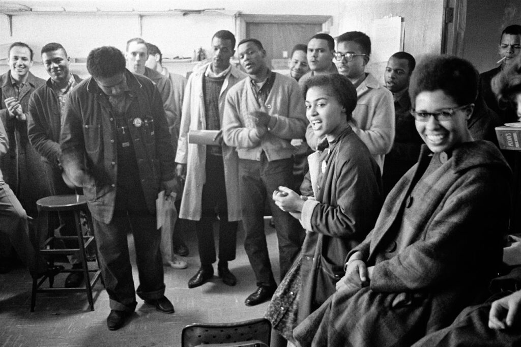 SNCC staff singing a freedom Song in SNCC’s national office  in Atlanta in 1963. They are wearing coats because there was no heat. Left to right: Mike Sayer, MacArthur Cotton, James Forman, Rick Manning, Marion Barry, Lester McKinnie, Mike Thelwell, Lawrence Guyot, Eric Jones, John Lewis (behind Jones), Julian Bond (far right rear with cigarette), Judy Richardson, Jean Wheeler. Photo credit: Danny Lyon, SNCC, courtesy SNCC Legacy Project